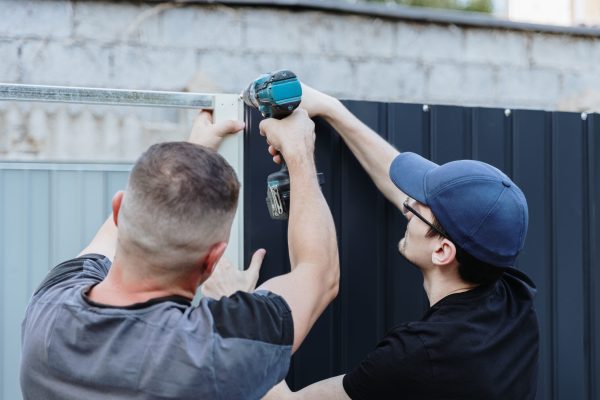 A mature Caucasian craftsman and his young assistant use a drill, each helping the other, to tighten a bolt while assembling a metal shed in the backyard on a summer day. Close-up side view. Conceptual metal shed assembly, teamwork, using technology.